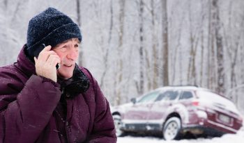 man on cellphone in snowstorm with car in snow bank on the side of the road