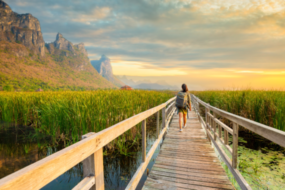 Asian hikers carry heavy backpacking on a small Pavilion outdoor hiking path on a wooden bridge in a swamp with meadows with a blue mountain background. Khao Sam Roi Yot National Park